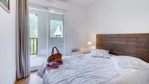 Une chambre à coucher comprenant un lit, une table de chevet, une lampe, une fenêtre avec des rideaux et une porte-fenêtre coulissante donnant sur un espace extérieur.