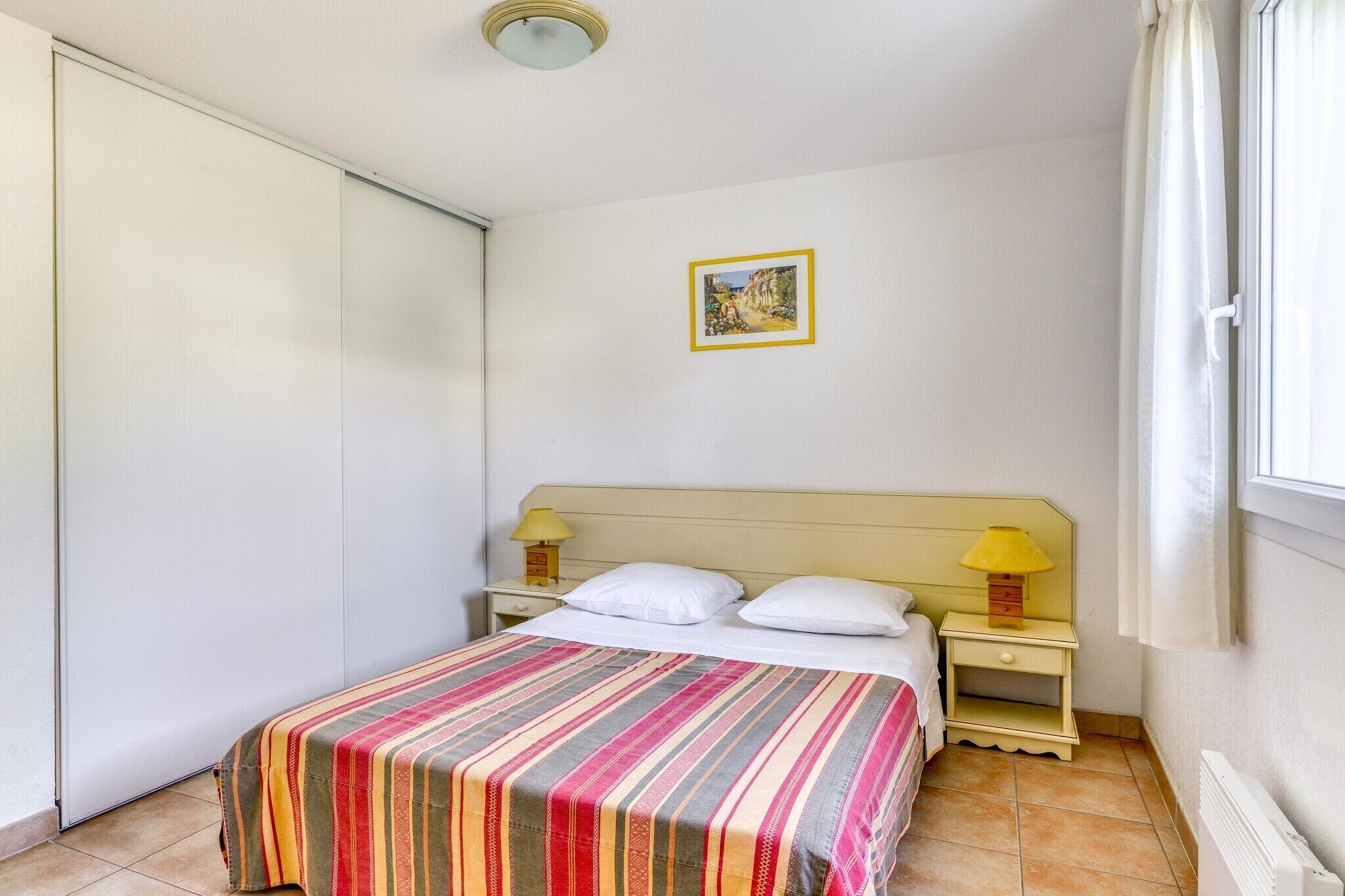 Une chambre à coucher avec un lit, deux tables de chevet avec des lampes, une porte, une fenêtre avec des rideaux et un tableau au mur.