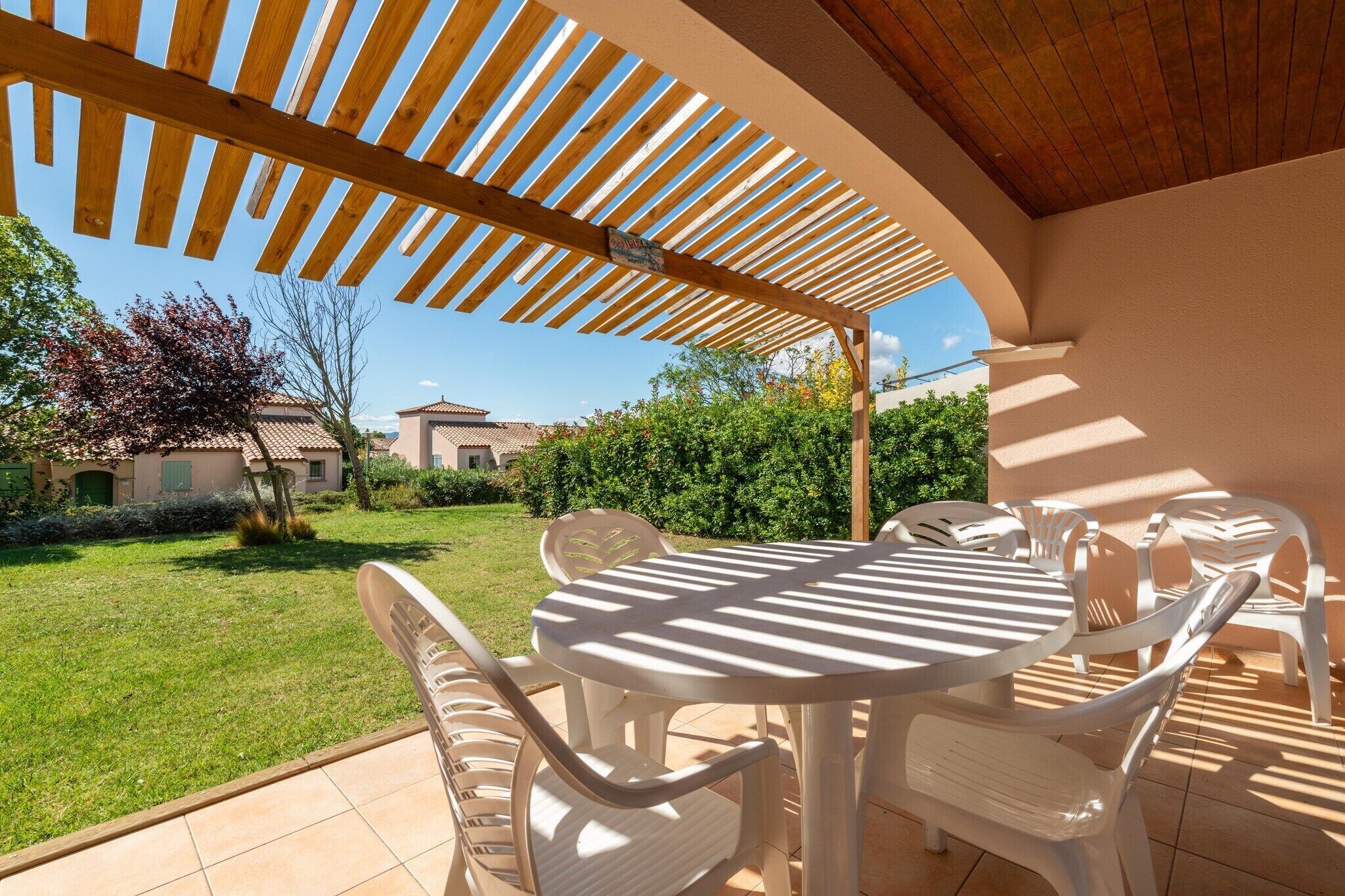 An outdoor patio with a round table and white plastic chairs, a wooden pergola overhead, and a view of a garden and buildings in the background.