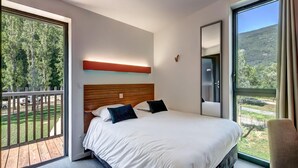 Ein modernes Schlafzimmer mit einem großen Bett, einem Holz-Kopfende, einem Balkon mit Blick auf Bäume und einem großen Fenster mit Blick auf Hügel.