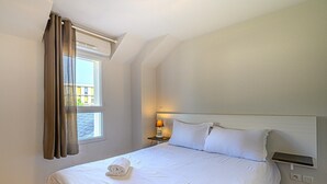 A hotel room with a bed, a nightstand with a lamp, a window with curtains, and a view of buildings outside. - Comfortable House Stay for 6 in Plescop (Plescop)