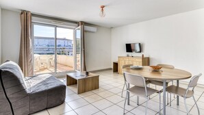 A modern living room with a sofa, a dining table with chairs, a TV, and a balcony with a view. - AC Apartment with Pool View for 4 (Le Grau-du-Roi)