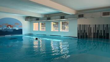 Una piscina interior con agua cristalina, un mural de montañas y ventanas amplias.