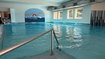 Piscina interior con vista a un mural de montaña, ventanas amplias y escalera metálica.