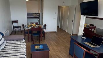 Une chambre d’hôtel compacte comprenant un lit, un bureau avec un ordinateur portable, un coin repas avec des chaises et un téléviseur fixé au mur.
