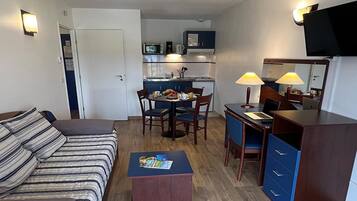 Un espacio habitable compacto con sofá, una pequeña mesa de comedor, kitchenette, escritorio con lámpara y televisor montado en la pared.