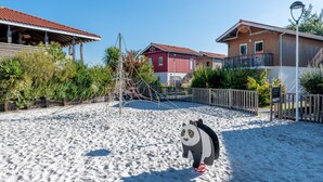 Zona de playa con una escultura de arena de un panda, edificios de madera y un parque infantil cercado.