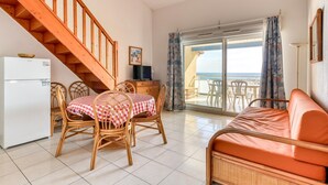 Ein Zimmer mit Esstisch, Stühlen, einer Couch, einer Holztreppe, einem Kühlschrank, einem Fernseher und Meerblick.