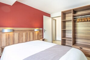 Ein Hotelzimmer mit Bett, Holz-Kopfteil, Kleiderschrank mit Platz zum Aufhängen und einer Tür zu einem anderen Raum.