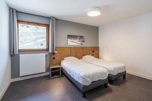 A bedroom with two single beds, a window with curtains, a radiator, a small wooden shelf, and a picture on the wall. - 6-Guest Alpine Rental with Balcony (AURIS-EN-OISANS)