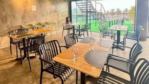 Un restaurante con mesas y sillas, vista a una escalera y follaje en el exterior, y una pared de concreto.