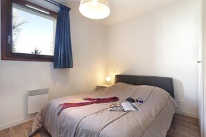 Un dormitori amb llit, finestra amb cortines, lampadeta de nit i radiador.