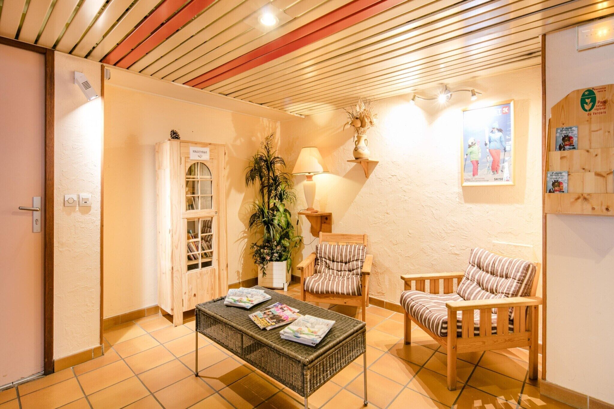 Une pièce comprenant un meuble en bois, une plante en pot, un fauteuil à rayures, une table en osier avec des magazines et une lampe fixée au mur.