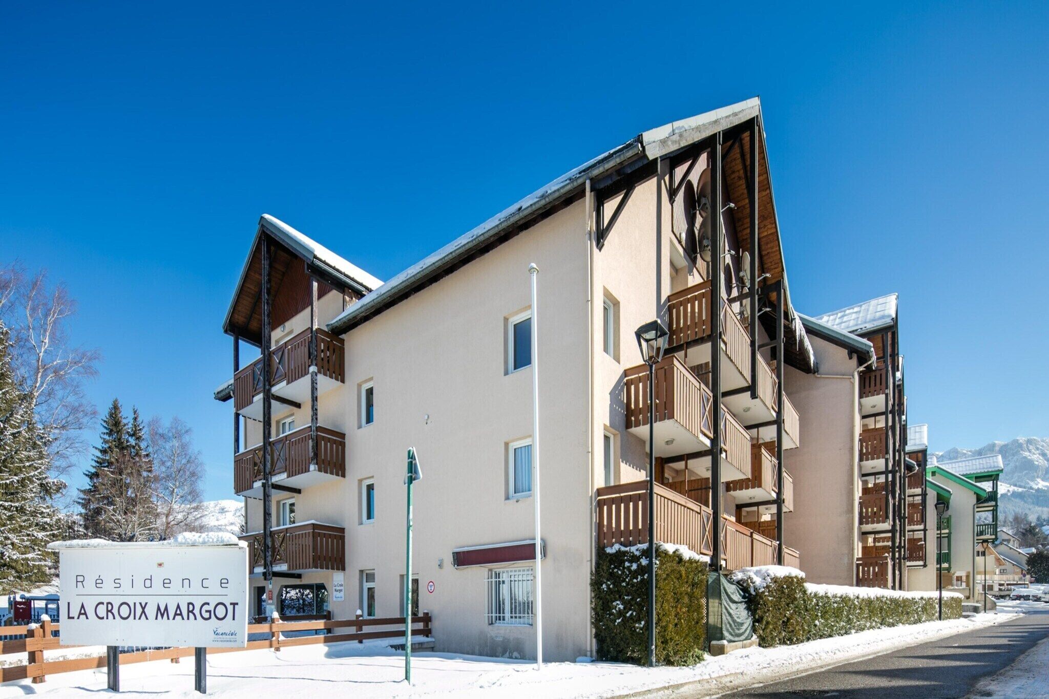 Un immeuble à plusieurs étages doté de balcons, une enseigne indiquant « Résidence LA CROIX MARGOT », et un paysage enneigé.