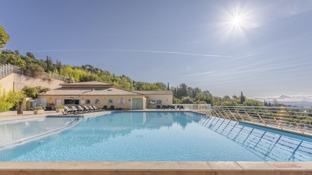 En swimmingpool med en solupplyst himmel, en byggnad med ett tak täckt av takpannor och en sluttning i bakgrunden.