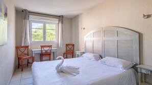 Quarto de hotel com cama, escrivaninha, duas cadeiras, janela com cortinas e luminária na parede.