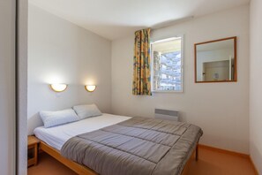 A single bed with a headboard, two wall-mounted lights, a window with curtains, and a mirror on the wall. - Alpine Base in Auris-en-Oisans (Germ)