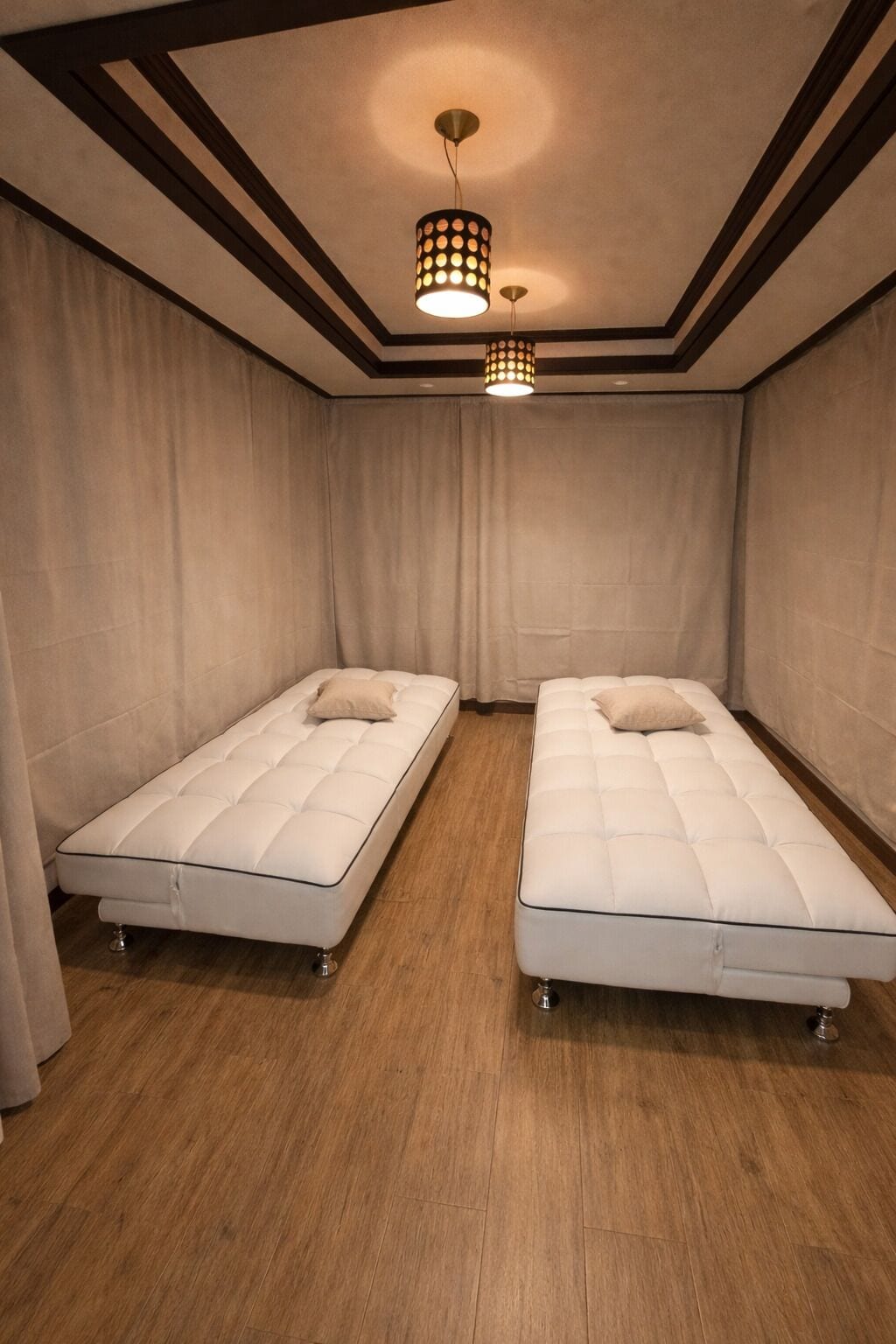 A room with two beds, wooden flooring, and a ceiling light fixture.