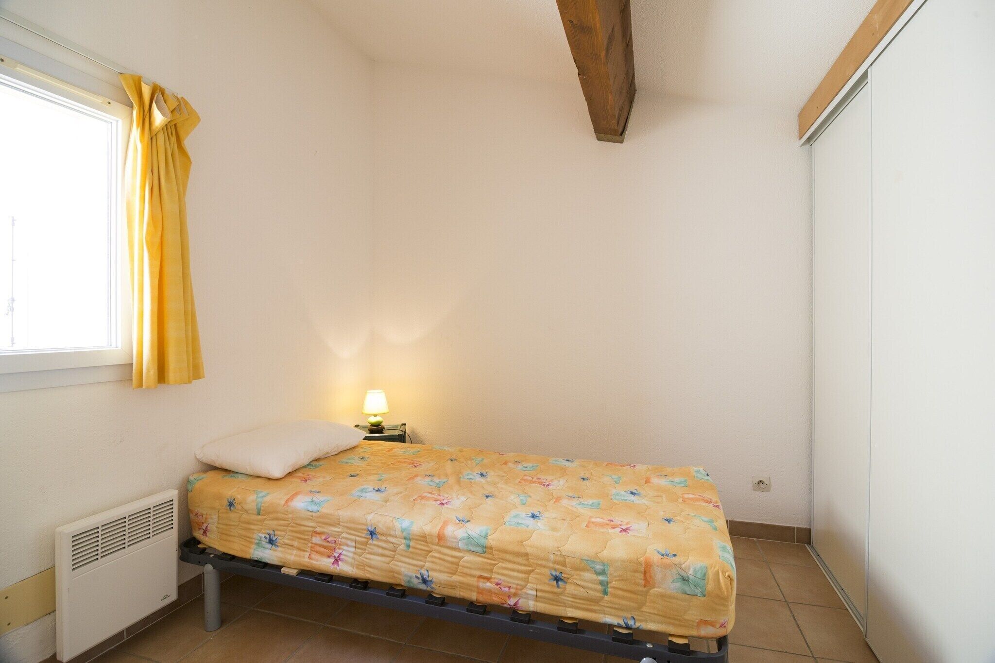 A single bed with a floral bedspread, a bedside lamp, and a window with yellow curtains.