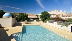 Una piscina con bordo piastrellato, circondata da edifici con tetti in terracotta e un cielo azzurro limpido.