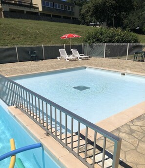Ein rechteckiger Swimmingpool mit einem Metallzaun, umgeben von einer gepflasterten Liegefläche und einer Rasenfläche mit Liegestühlen und einem Sonnenschirm.