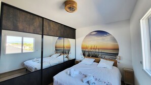 Een slaapkamer met een bed, een grote spiegel, een wandlamp en uitzicht op het strand.