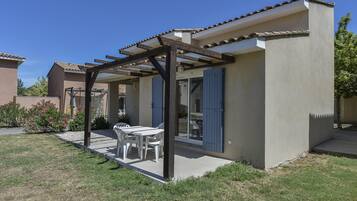 Un patio couvert avec une table et des chaises, une pergola et un bâtiment aux volets bleus.
