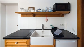 Une cuisine avec un plan de travail noir, un évier blanc, des placards en bois, un four à micro-ondes et une étagère où sont rangés des verres.