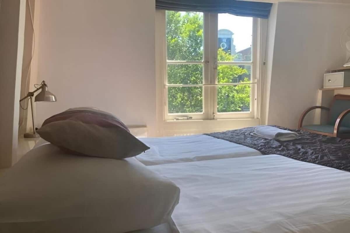 A hotel room with a bed, a window with a view of greenery, a desk with a lamp, and a chair.