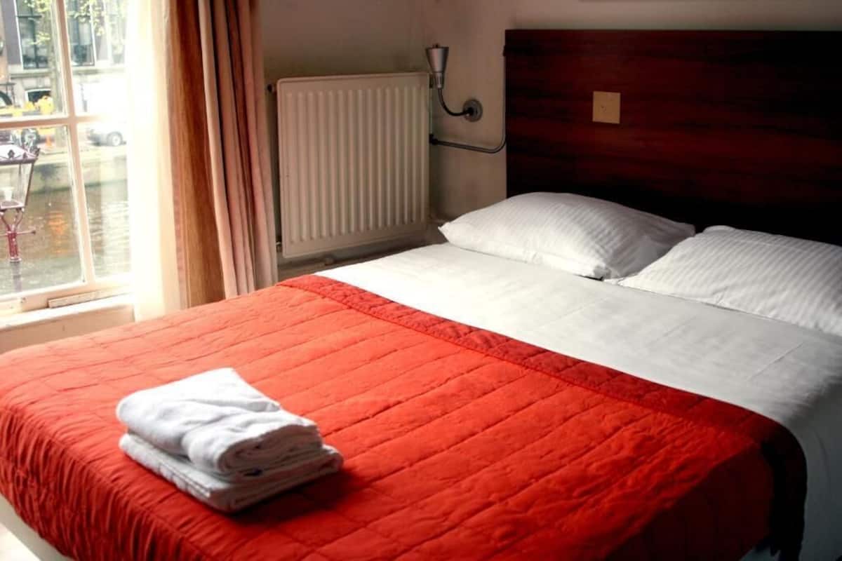 A bed with a red quilt, white pillows, and a folded white towel on the side.