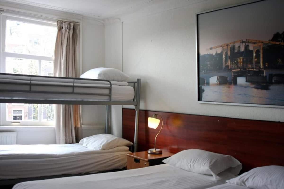 A hotel room with a bunk bed, a bedside table with a lamp, a window with curtains, and a cityscape painting on the wall.