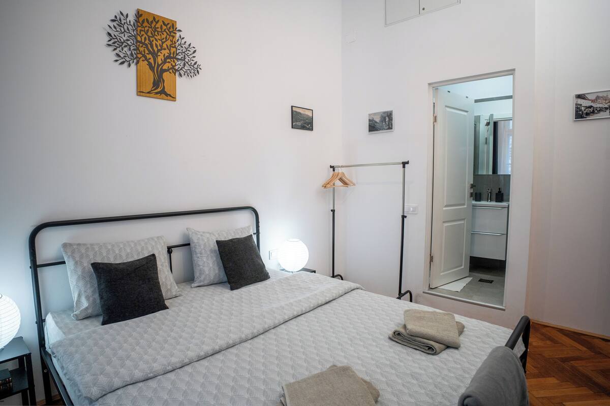 Un dormitorio con cama, almohadas, una mesita de noche, una lámpara colgante y una puerta que da a un baño.