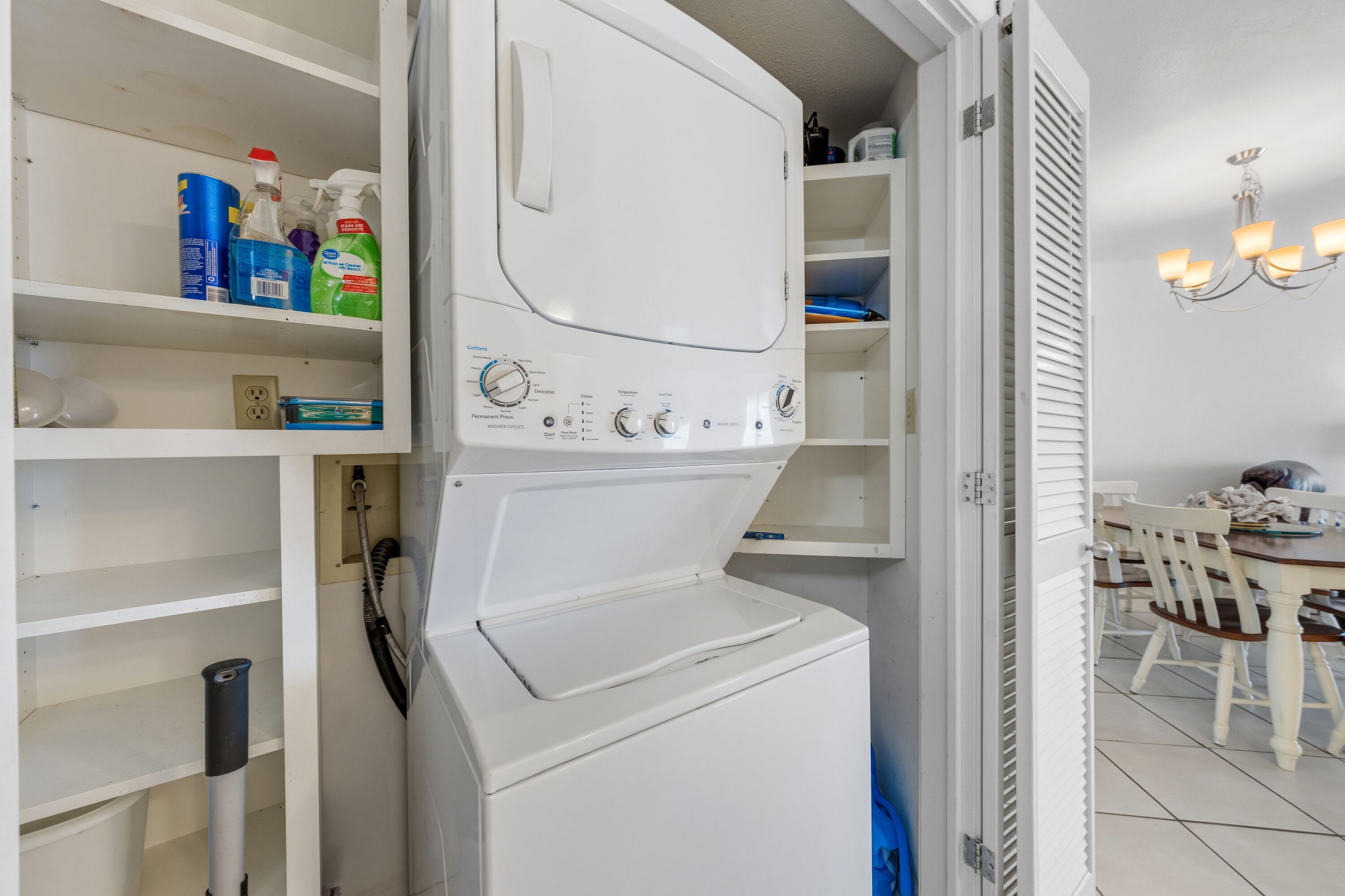 A laundry room with a white washing machine, shelves with cleaning supplies, and a dining area with a chandelier.