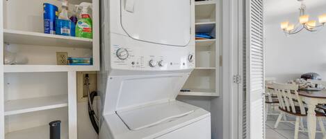 A laundry room with a white washing machine, shelves with cleaning supplies, and a dining area with a chandelier.