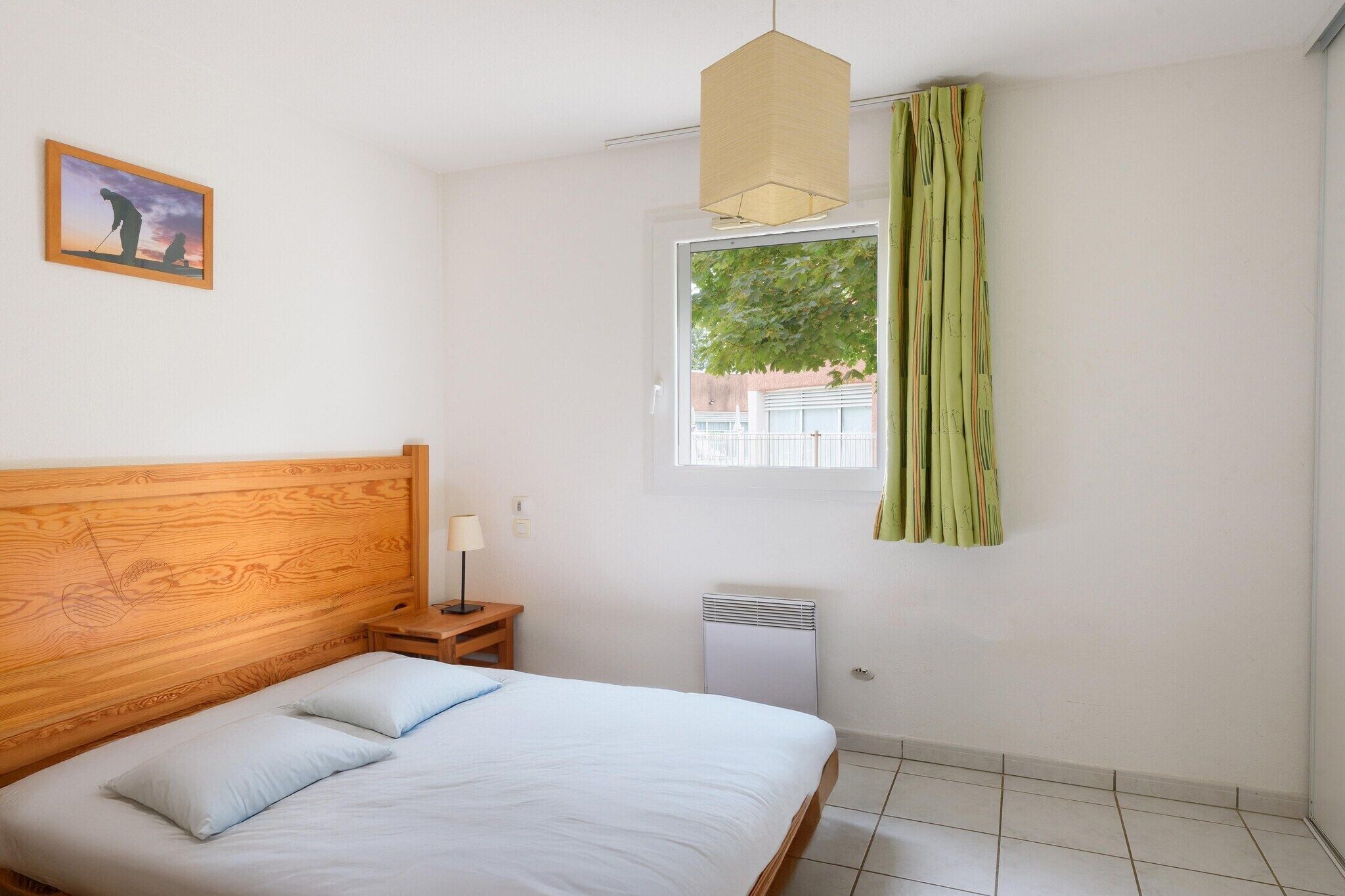 Une chambre à coucher avec une tête de lit en bois, un lit avec une literie blanche, une table de chevet avec une lampe, une fenêtre donnant sur un paysage verdoyant et un radiateur.