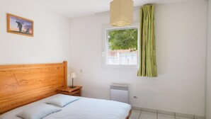 Un dormitorio con cabecera de madera, cama con ropa de cama blanca, mesita de noche con lámpara, ventana con vista a la vegetación y un radiador.
