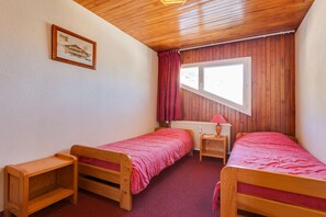 A small room with two wooden beds, a window with curtains, a radiator, and a framed picture on the wall.