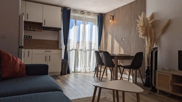 Ein modernes Wohnzimmer mit einer Couch, einem Esstisch mit Stühlen, einer Küchenzeile mit Schränken und einem großen Fenster mit Vorhängen.