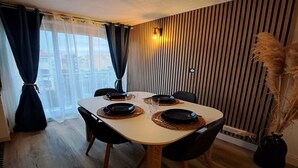 Ein Essbereich mit einem Tisch für vier Personen, Holzböden, einem Fenster mit Vorhängen und einer Wand mit vertikalen Holzleisten.