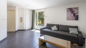 A modern living room with a grey sofa, a glass coffee table, and a large window with a view of greenery. - Apartment in Lacanau near beach (Lacanau)