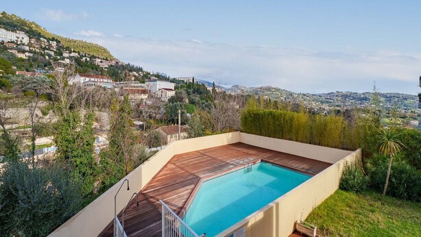Une piscine dotée d’une terrasse en bois, entourée de verdure et offrant une vue sur une ville située sur une colline.