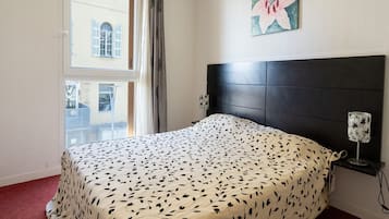 A bedroom with a bed, a headboard, a nightstand, a lamp, a window, and a painting.