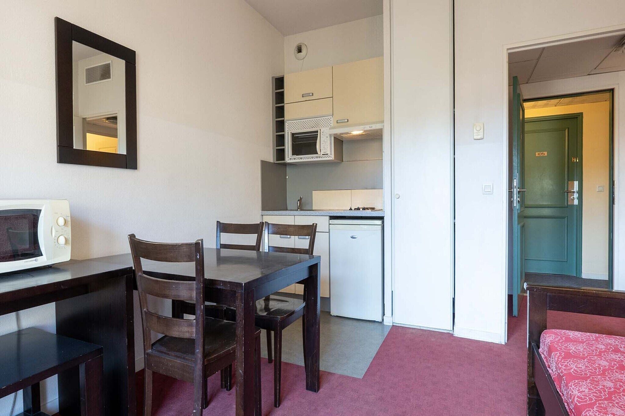 A compact room with a kitchenette, a dining table with chairs, a microwave, and a bed with a red bedspread.
