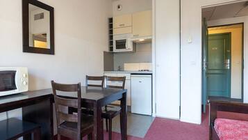 A compact room with a kitchenette, a dining table with chairs, a microwave, and a bed with a red bedspread.