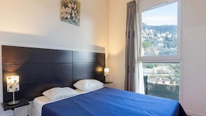 Hotelkamer met een bed, twee bedlampen, een schilderij aan de muur en een raam met uitzicht op een stad.