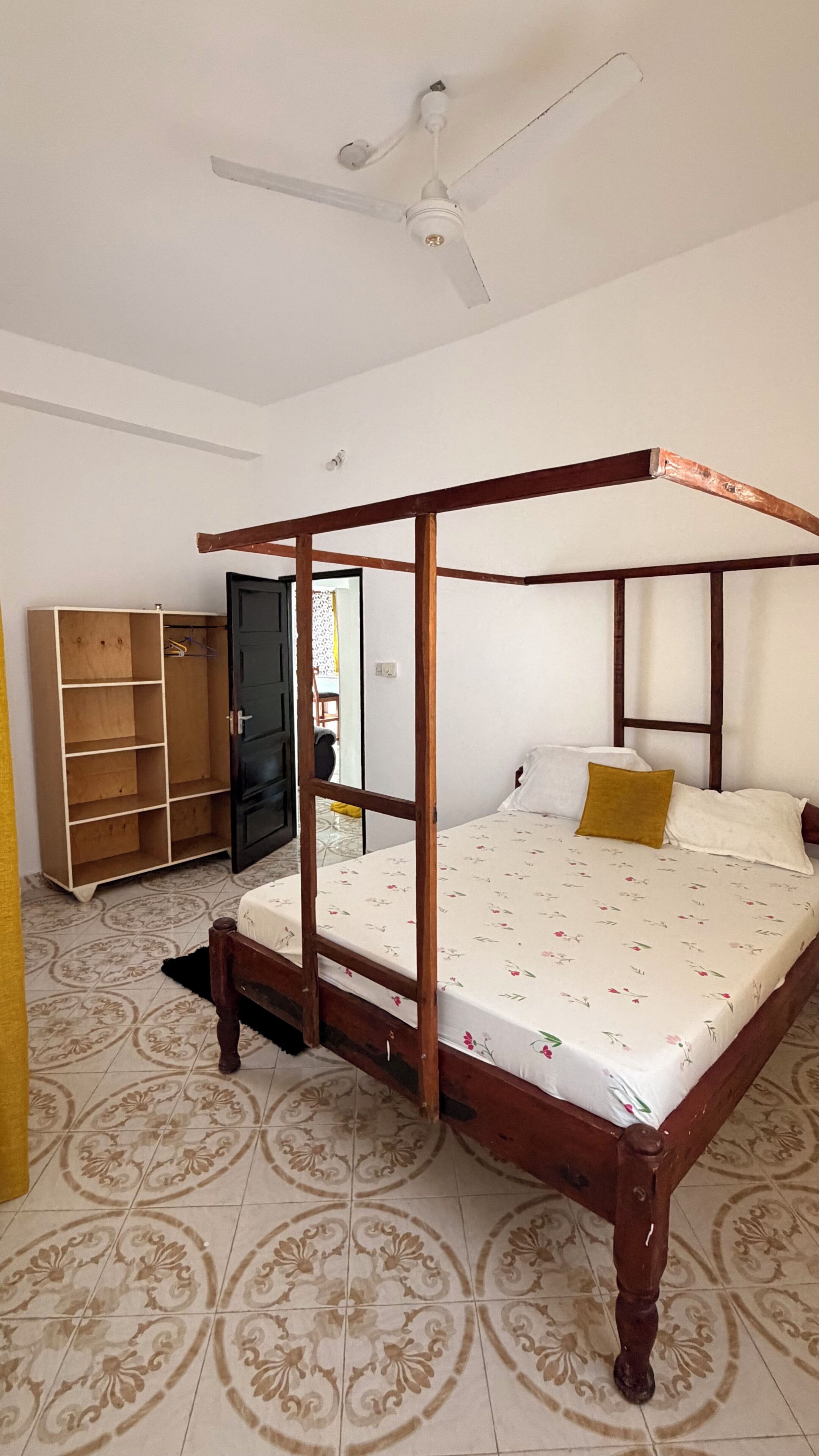 A four-poster bed with a wooden frame, a ceiling fan, and patterned floor tiles.