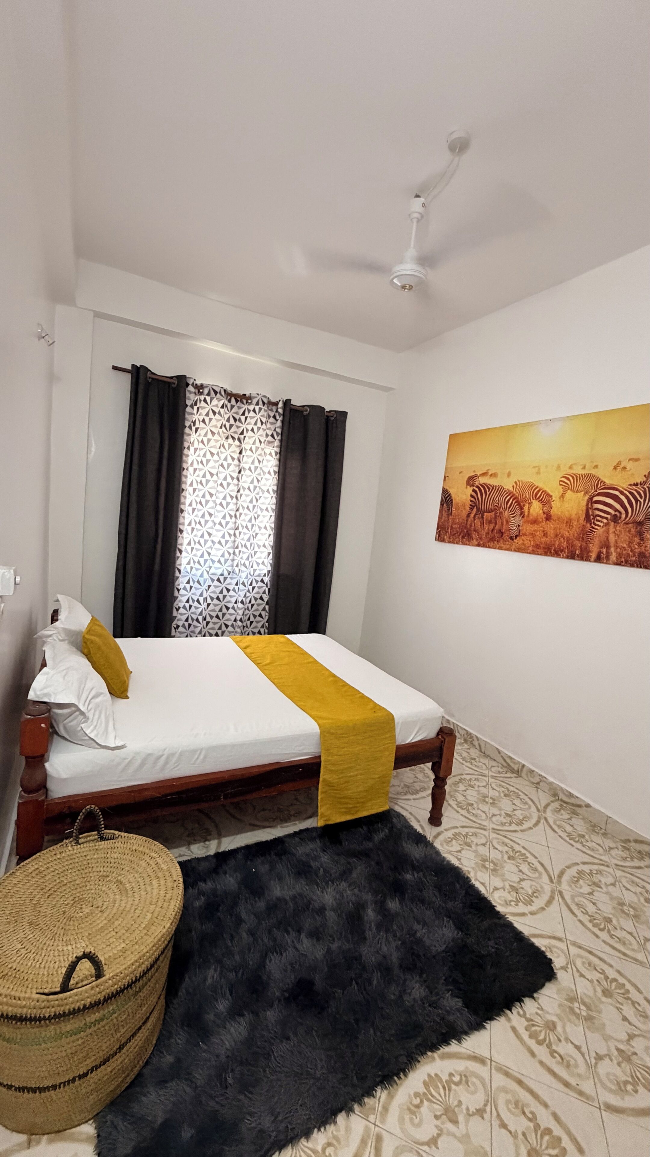 Ein Schlafzimmer mit einem Bett, einem Deckenventilator, einem Fenster mit Vorhängen, einem Zebra-Gemälde und einem gewebten Korb.
