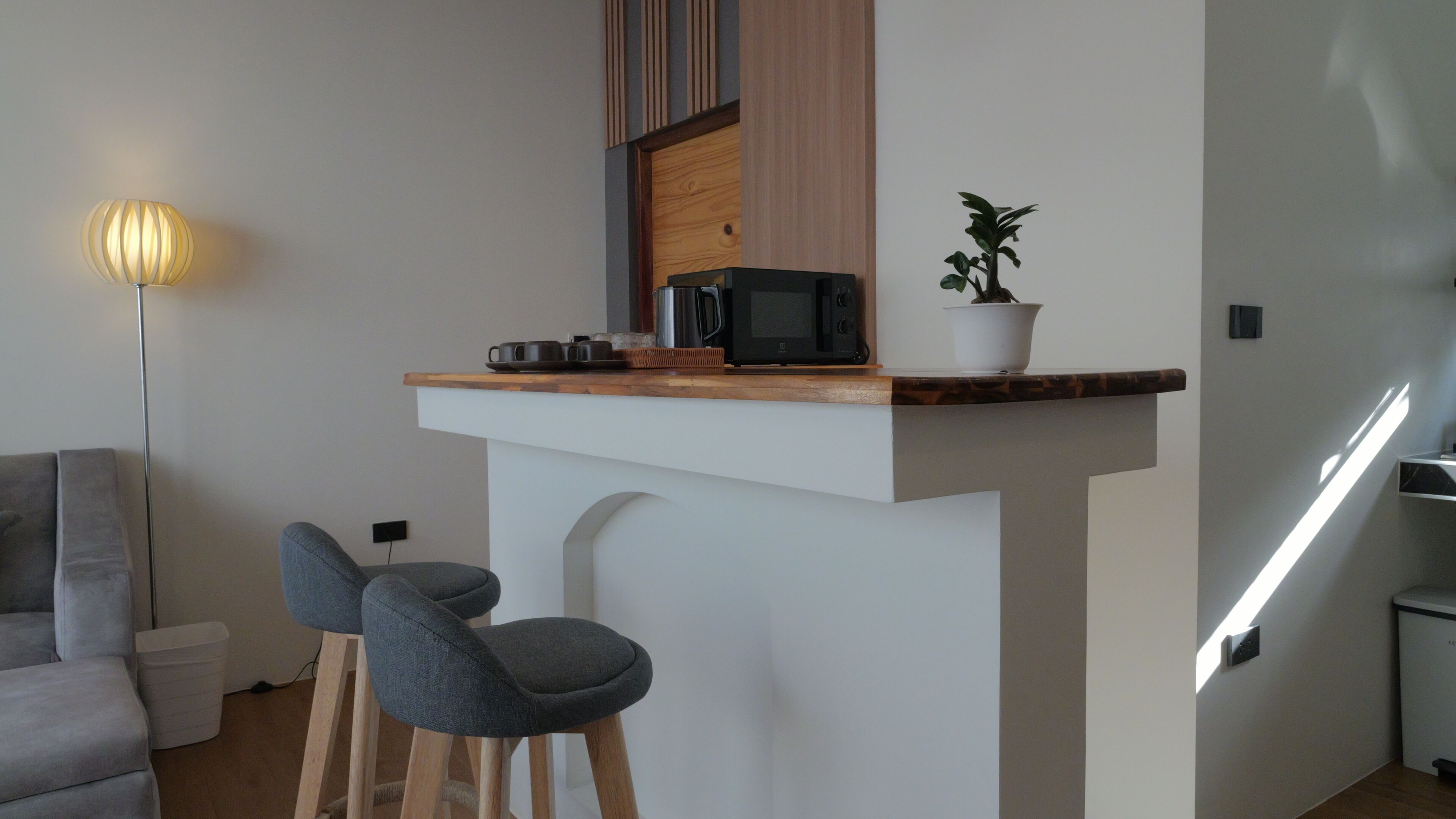Una cocina compacta con microondas, tostadora y planta en maceta sobre una encimera de madera. En la zona de estar contigua, hay dos taburetes tipo bar y un sofá gris.