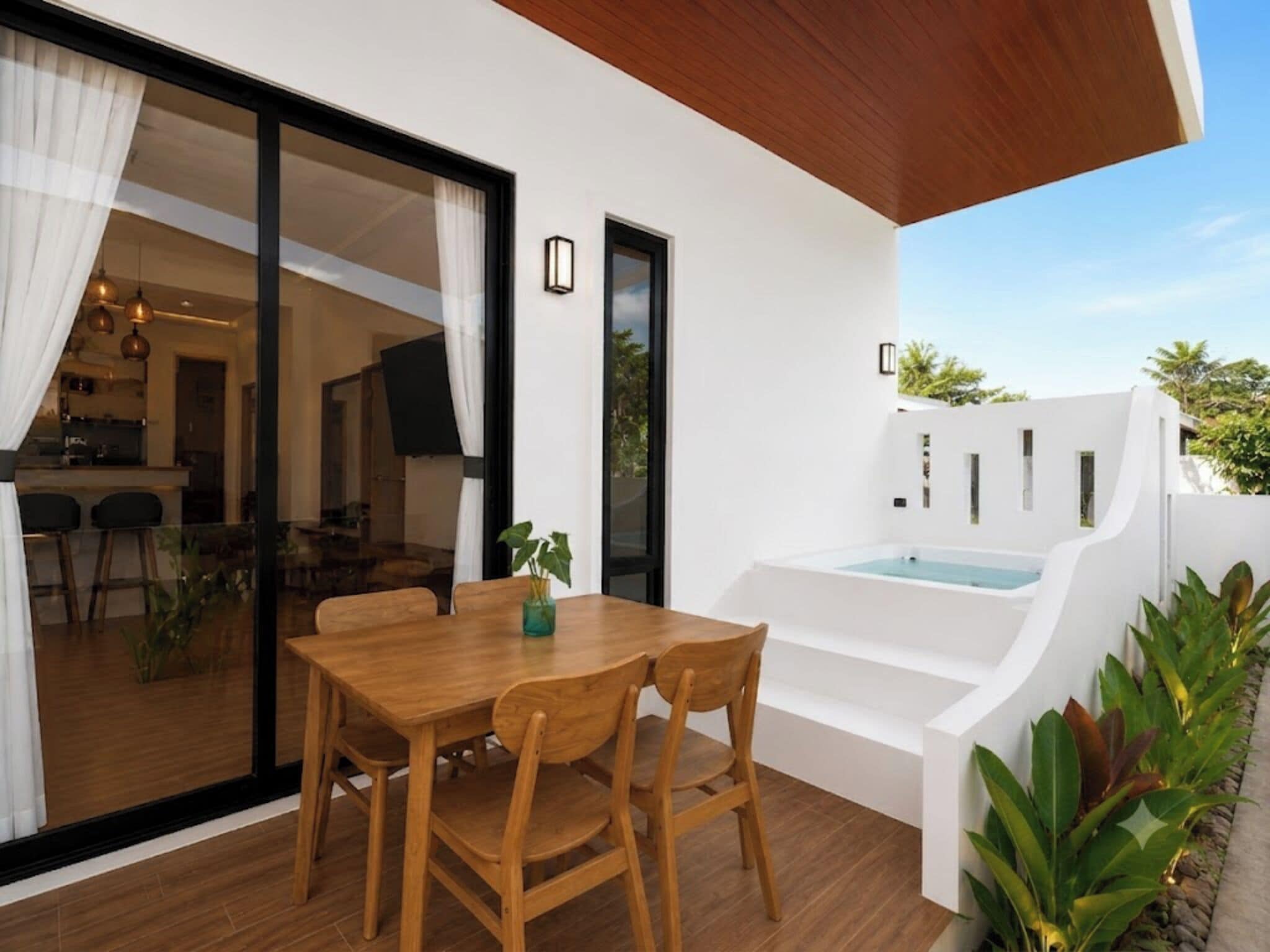 Un área de comedor exterior moderna con una mesa y sillas de madera, una puerta de cristal que da a una cocina y una bañera exterior blanca.