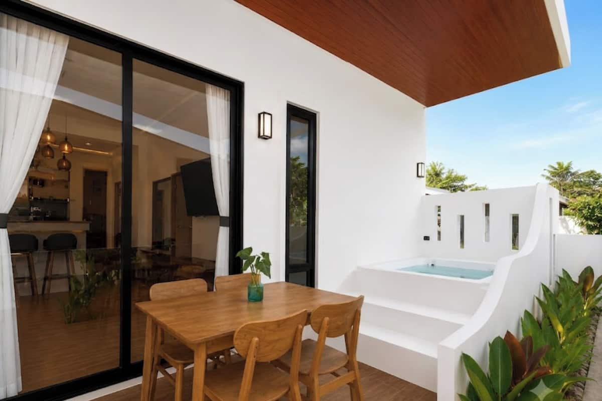 Un área de comedor exterior moderna con una mesa y sillas de madera, una puerta de cristal que da a una cocina y una bañera exterior blanca.
